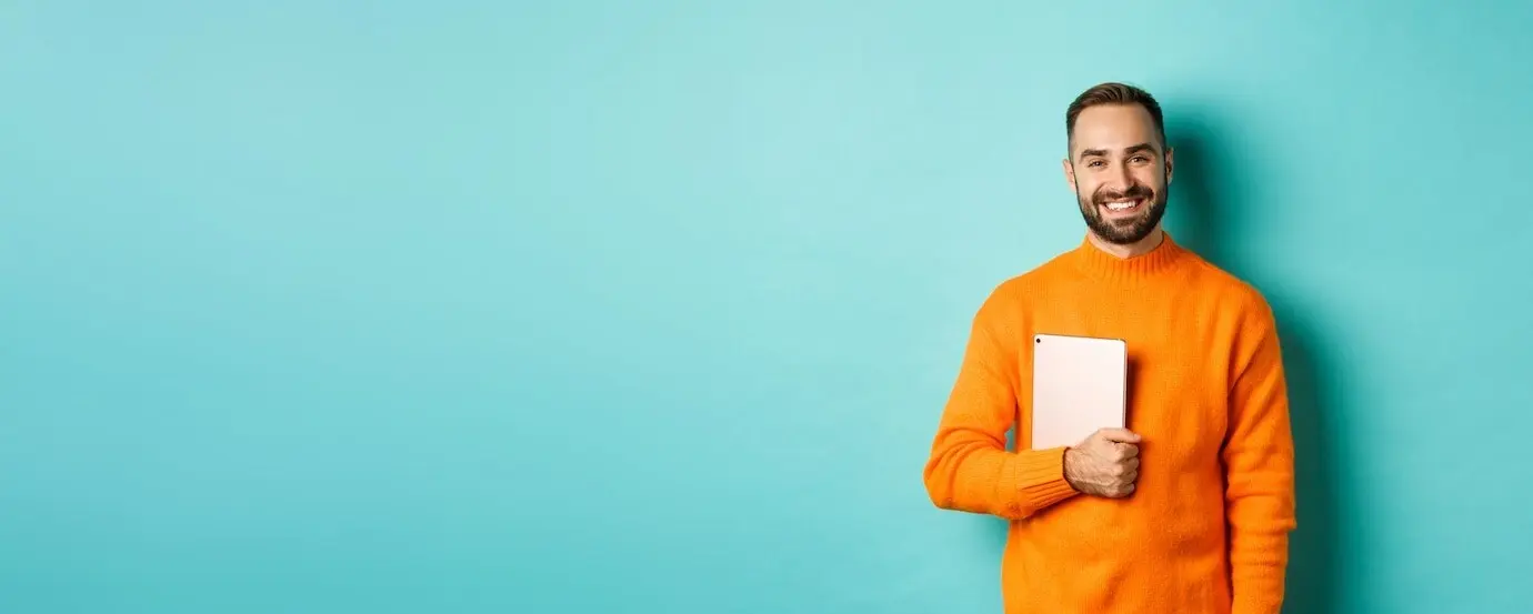 Gutaussehender freiberuflicher Mann, der einen Laptop hält und glücklich lächelnd vor einem hellblauen Hintergrund steht.